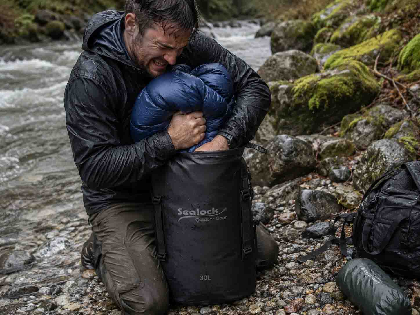Real gear test failure photo of a hiker struggling to compress an uncompressed synthetic sleeping bag inside a stiff 30L matte TPU dry bag on river rocks